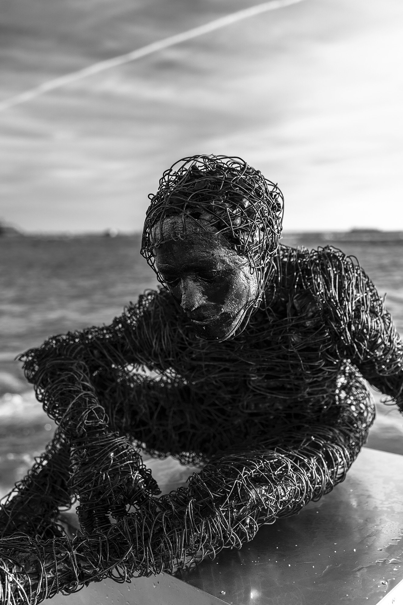 wire sculpure on a metal cube, standing from the sea in front of an orange sunset. Black and white image.