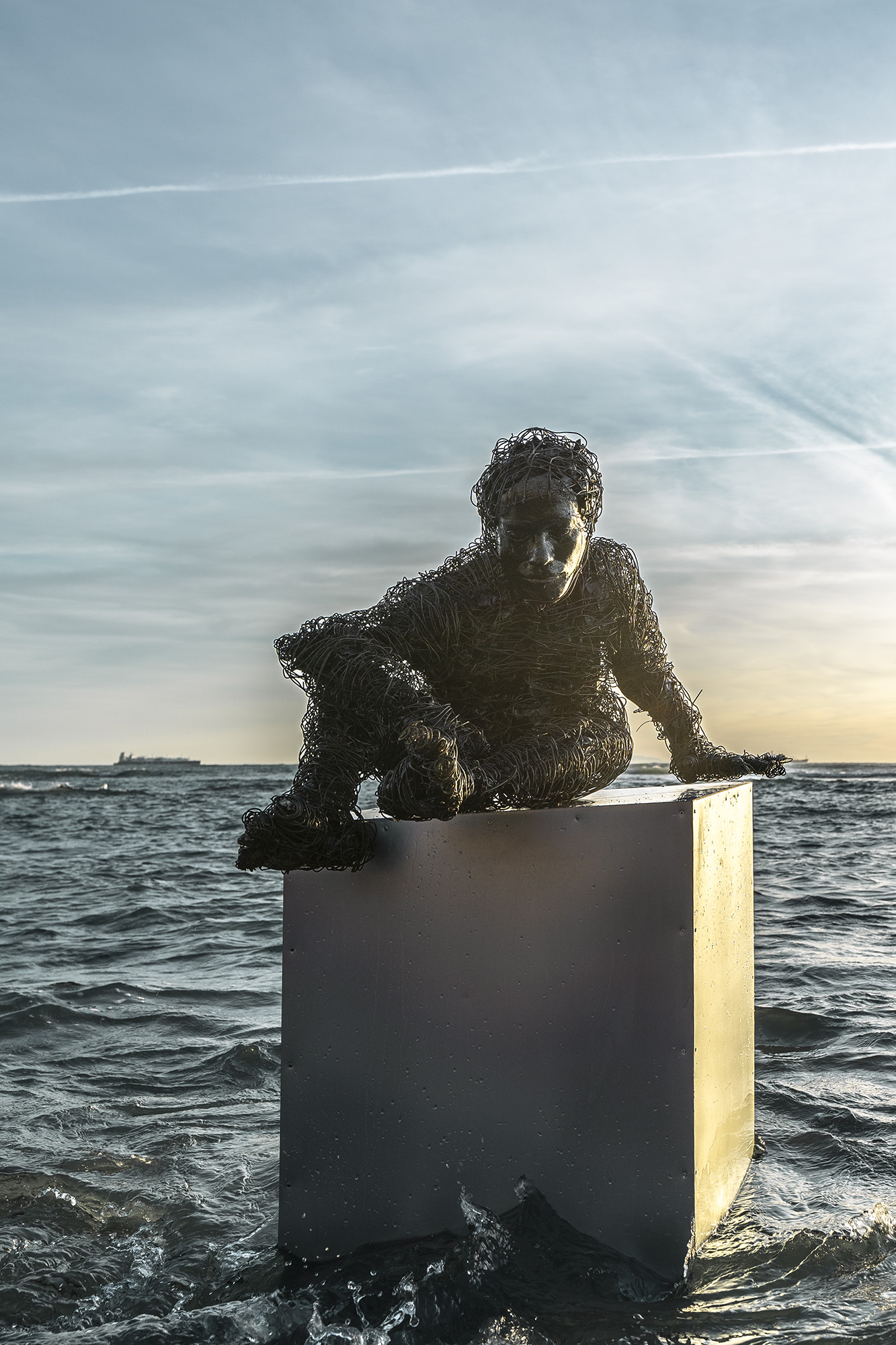 wire sculpure on a metal cube, standing from the sea in front of an orange sunset