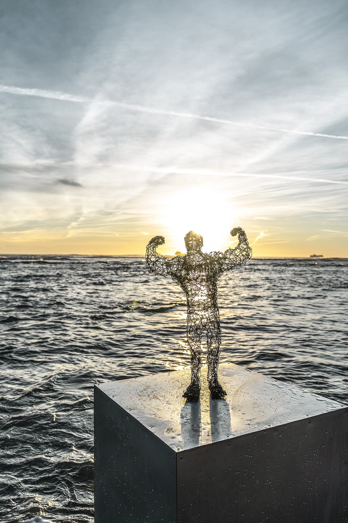 wire sculpure on a metal cube, standing from the sea in front of an orange sunset