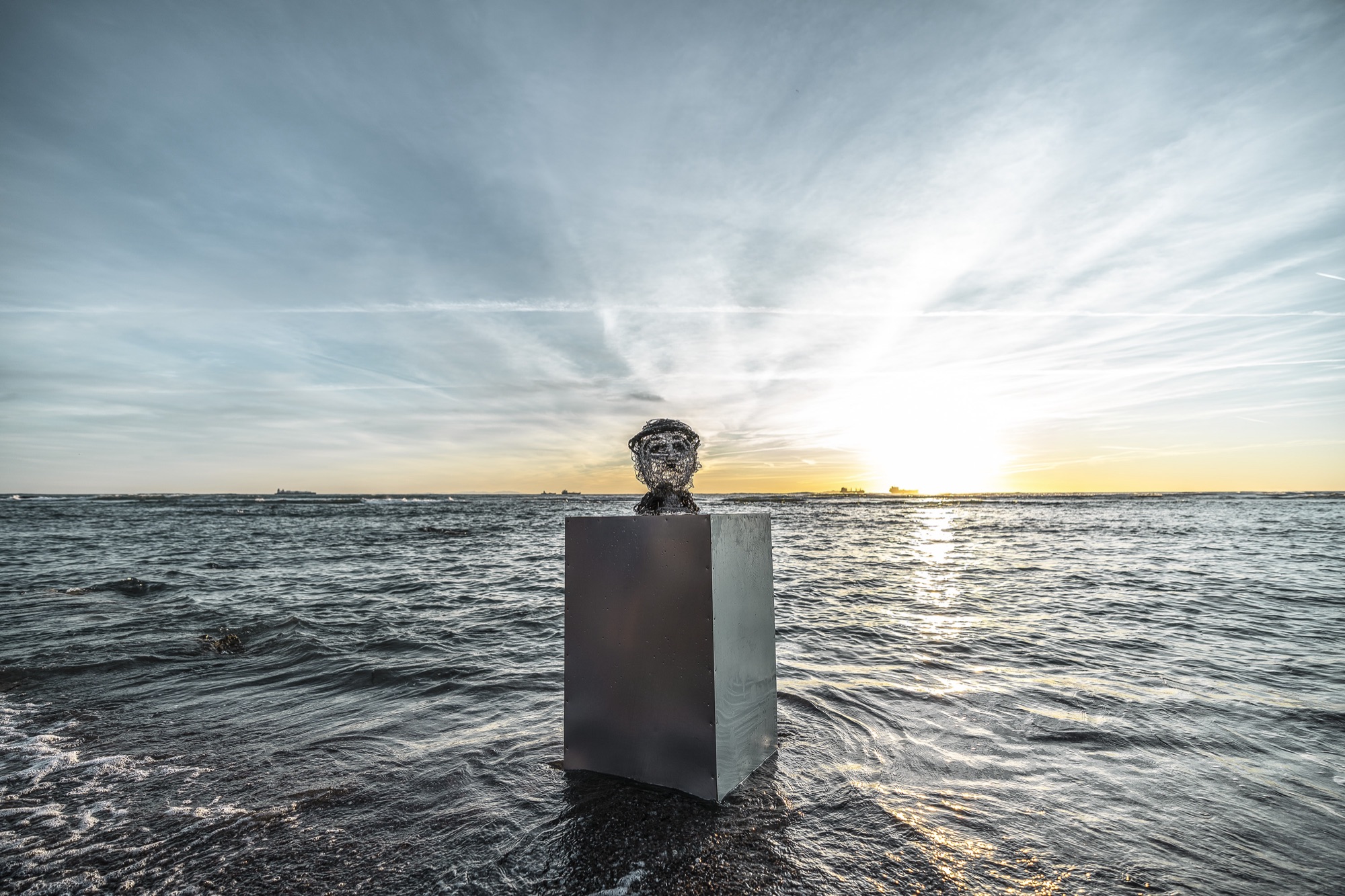 wire sculpure on a metal cube, standing from the sea in front of an orange sunset