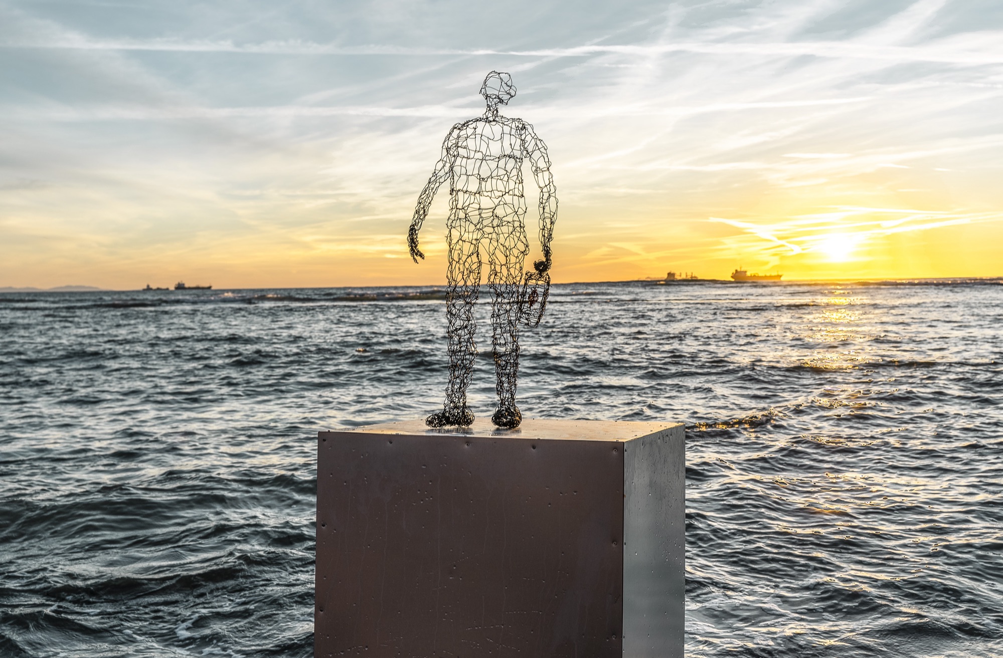 wire sculpure on a metal cube, standing from the sea in front of an orange sunset