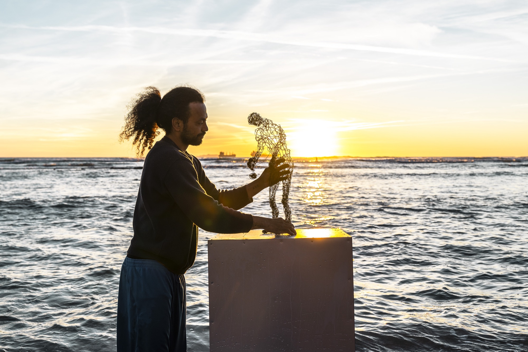 wire sculpure on a metal cube, standing from the sea in front of an orange sunset