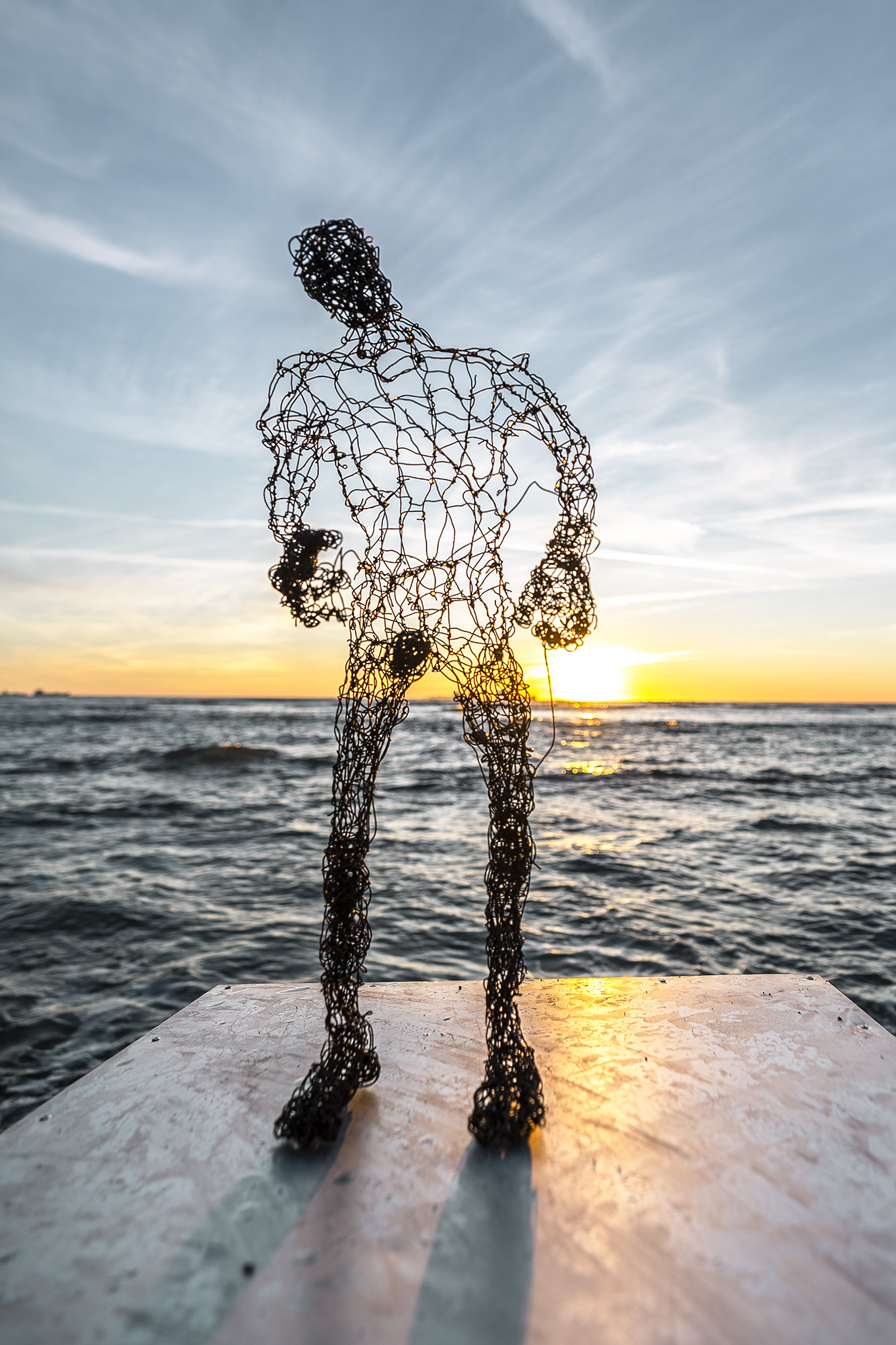 wire sculpure on a metal cube, standing from the sea in front of an orange sunset